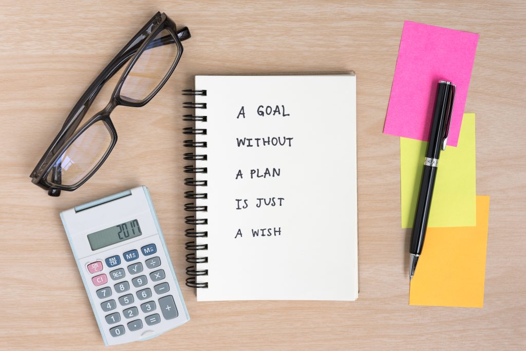 An eye glass, calculator, pen, colored sticky note and a notebook with the words- A goal without a plan is just a wish